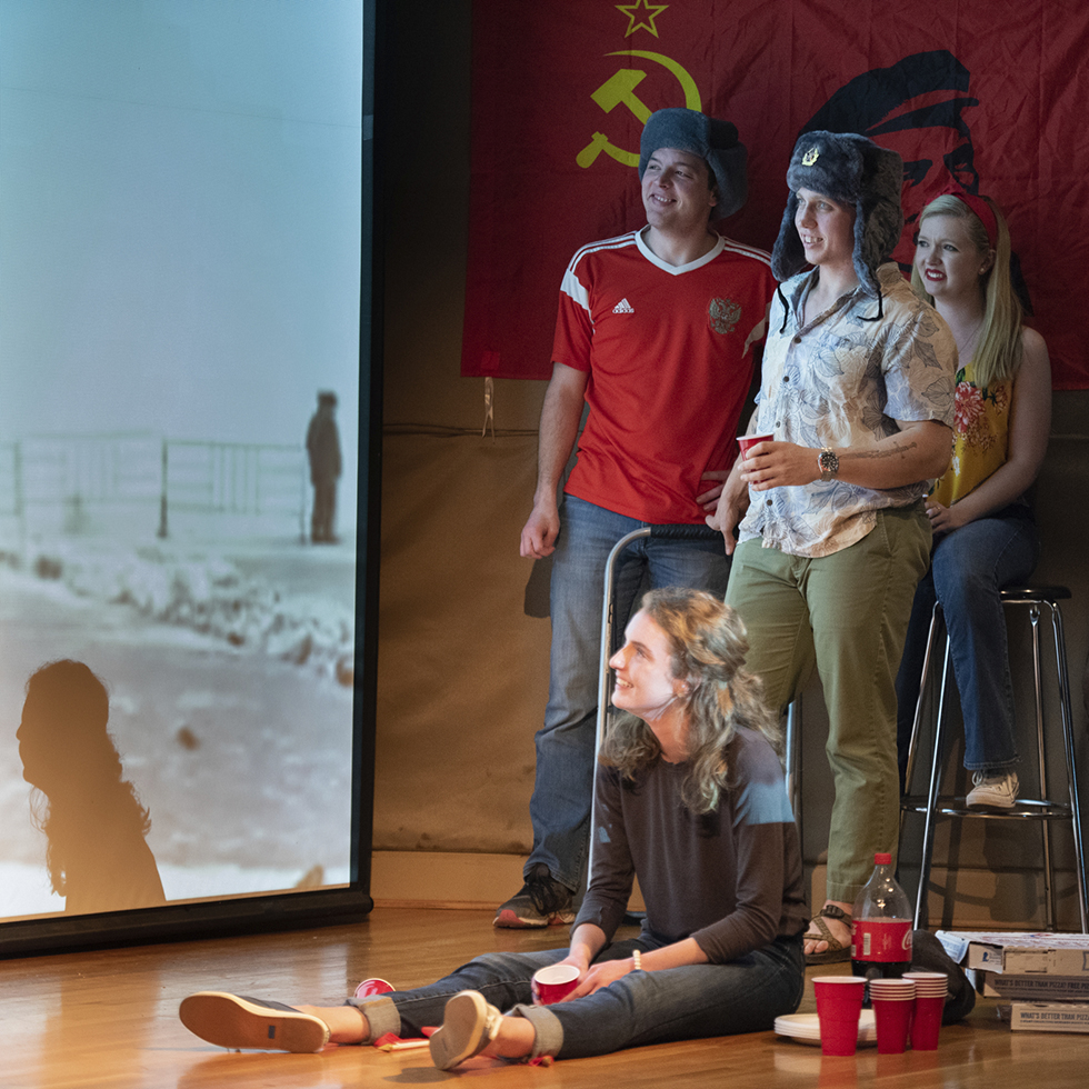 Students on a stage watch a video on a screen