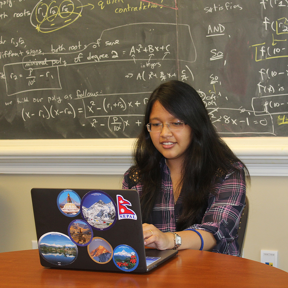 A student works at a laptop in front of a chalkboard displaying mathematical equations